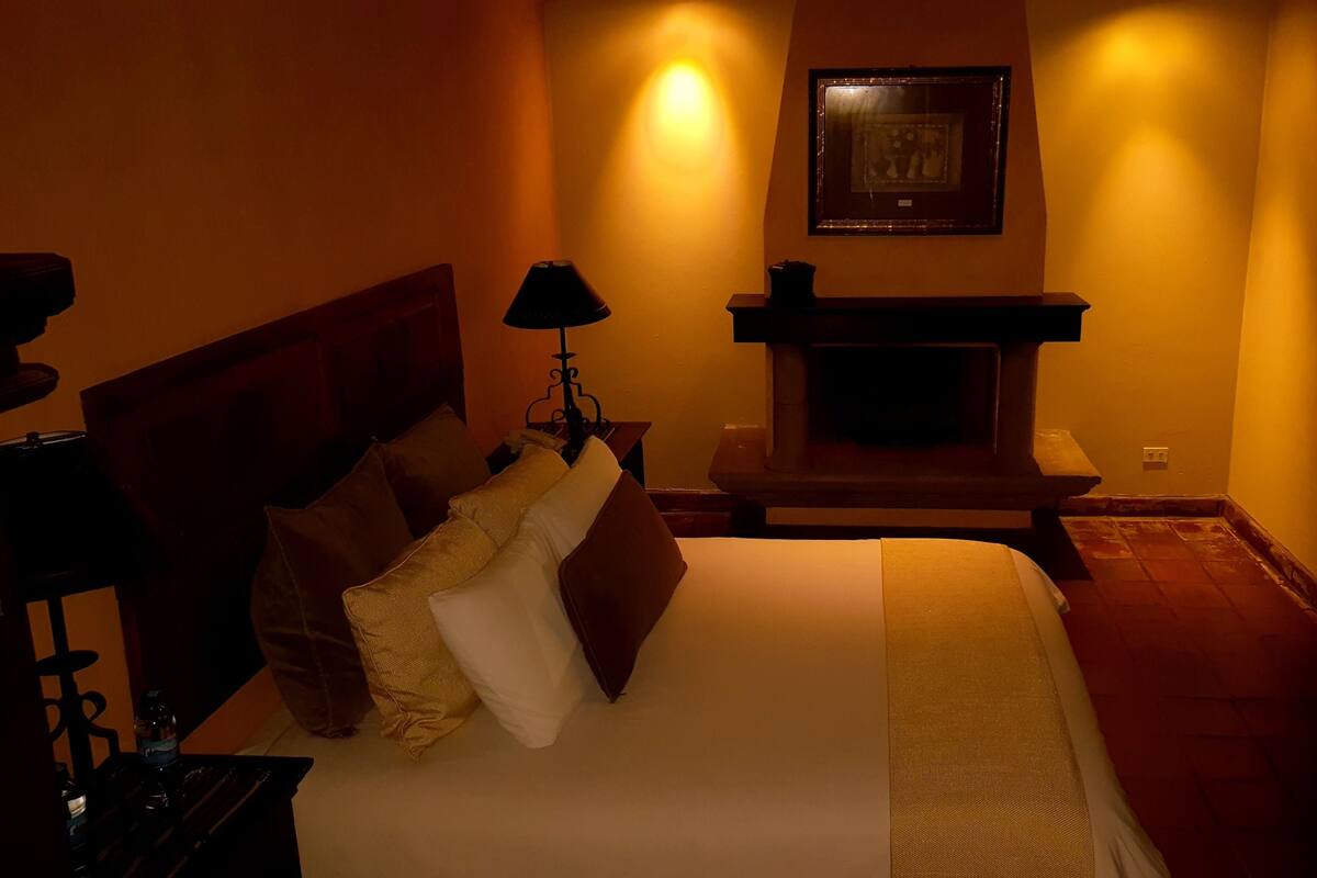 Una habitación de hotel con cama, mesita de noche, lámpara, chimenea y un cuadro enmarcado en la pared.