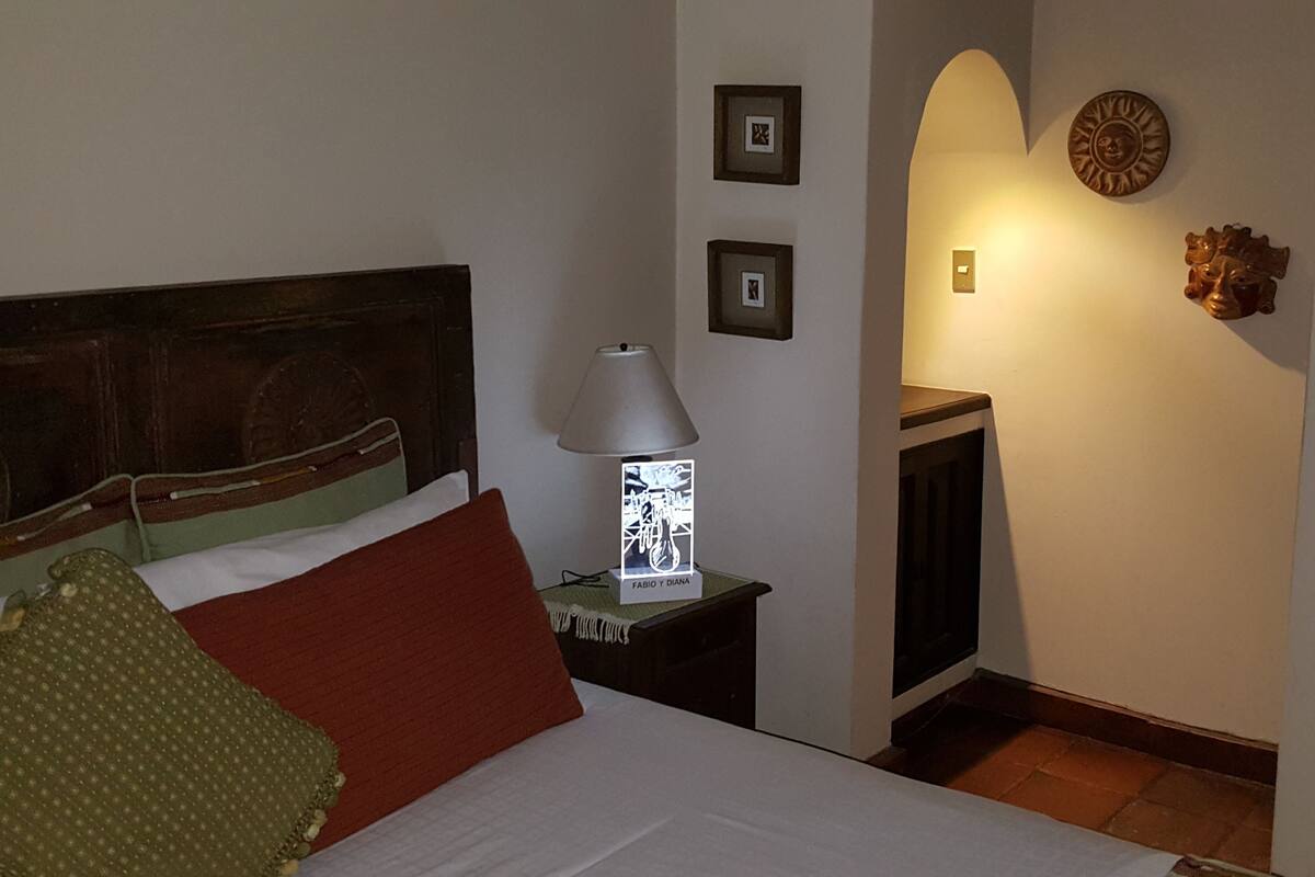 Un dormitorio con cabecera de madera, cama con almohadas rojas y verdes, mesita de noche con lámpara, y pared decorada con cuadros y un sol decorativo.