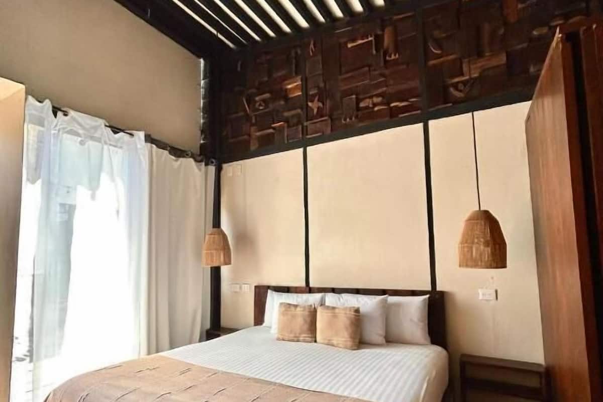 Un dormitorio con techo de madera, una cama con colcha color beige, dos lámparas de noche y una ventana con cortinas blancas.