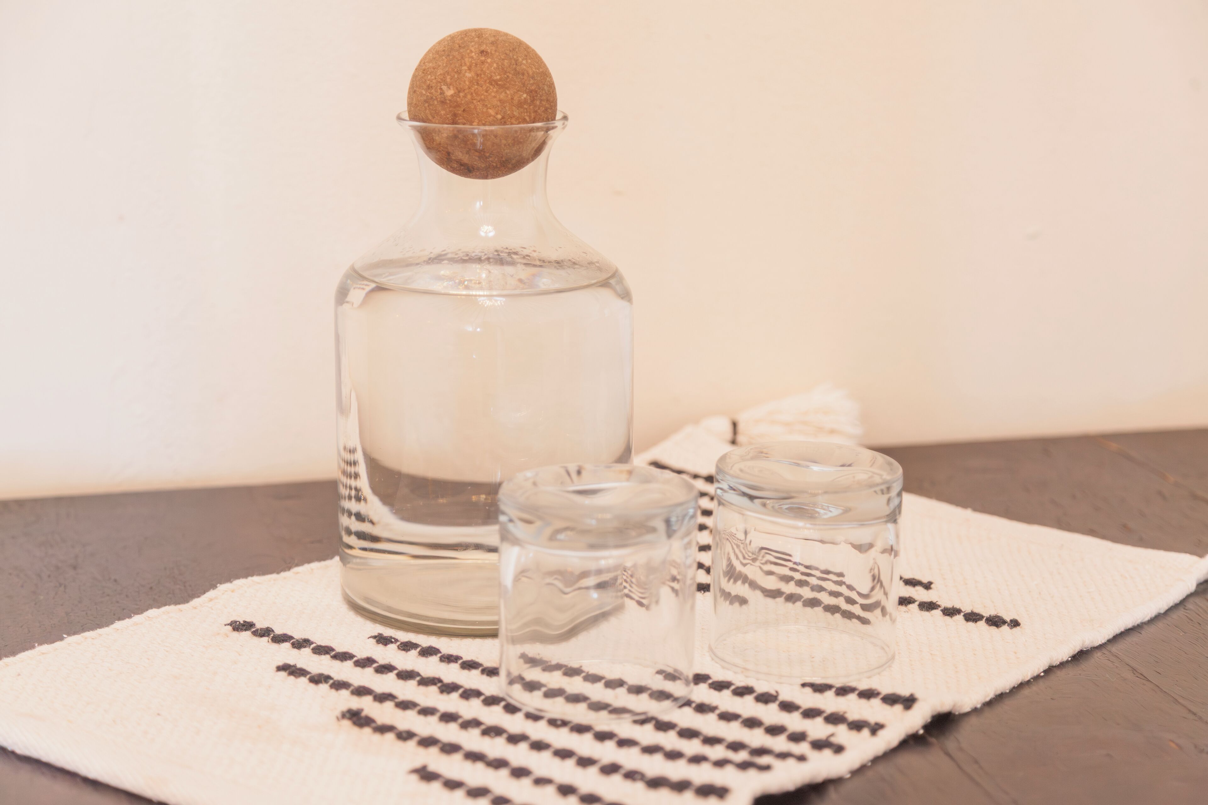 A clear glass bottle with a cork stopper, two empty glass cups, and a white cloth with black patterns on a wooden surface.