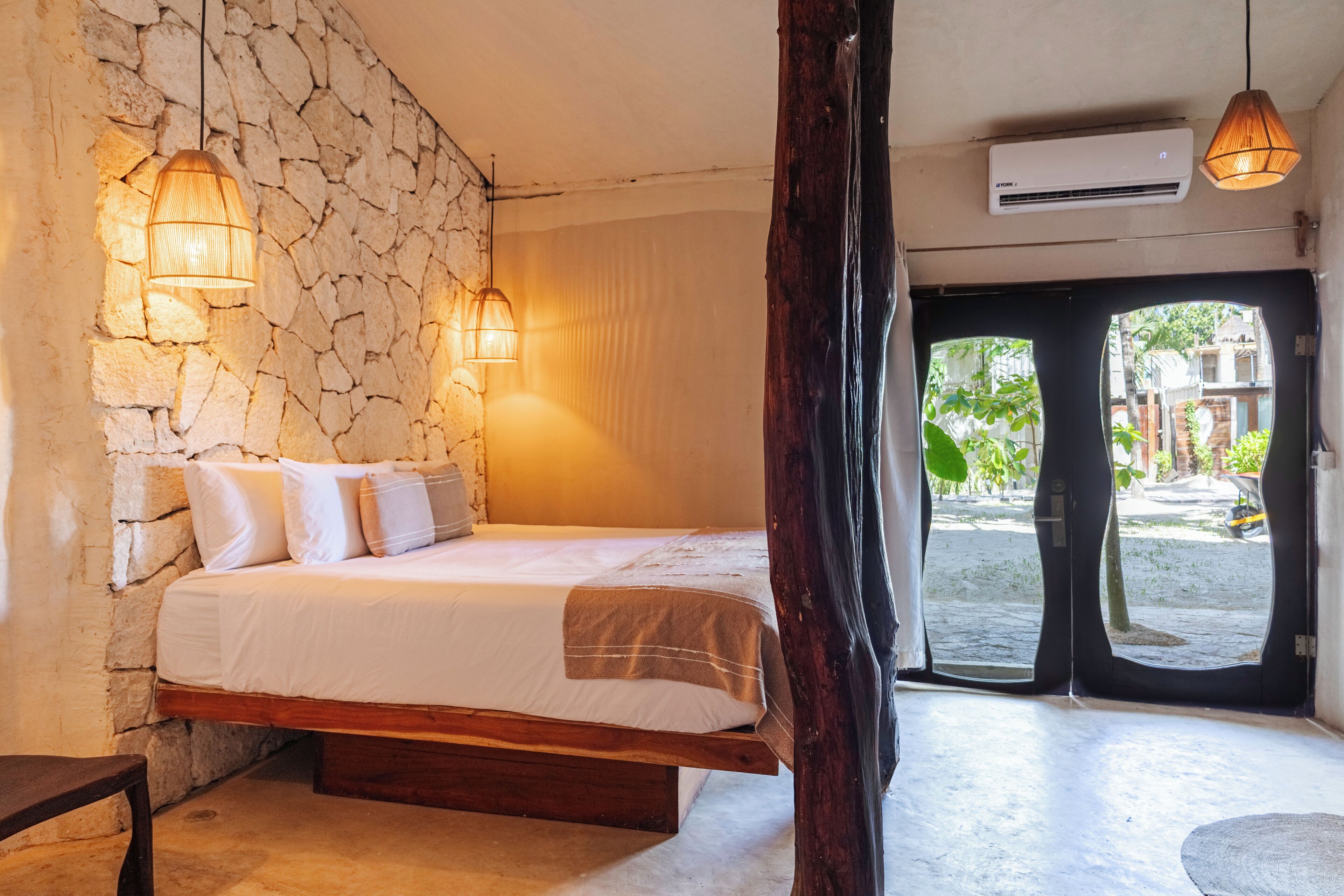 Un dormitorio con pared de piedra, cama con ropa de cama blanca, dos lámparas colgantes y un equipo de aire acondicionado.