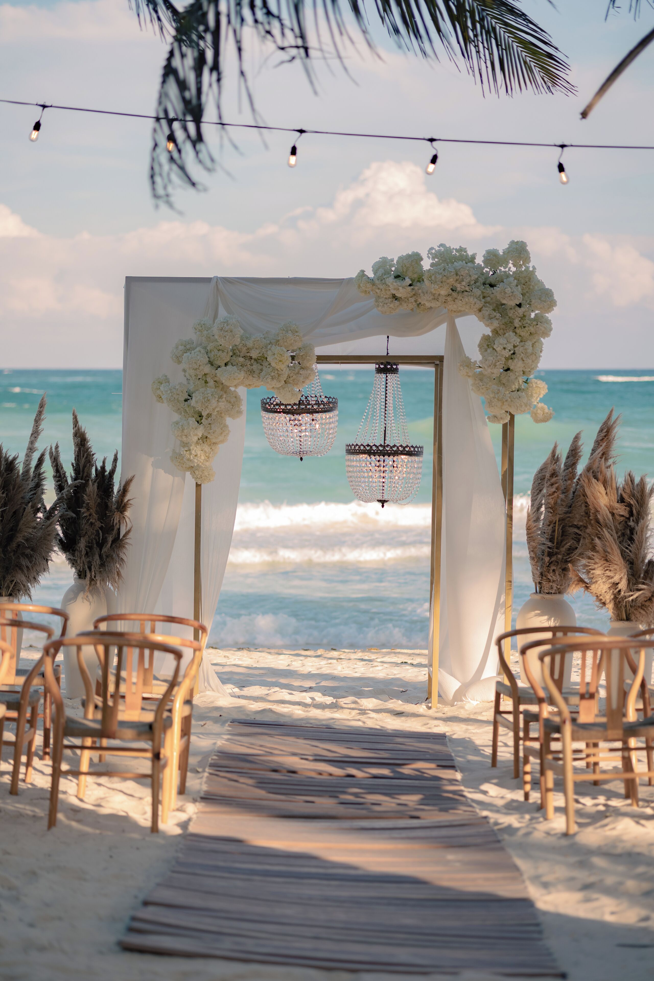 Decoración para una boda en la playa, con un arco blanco, sillas de madera y un camino que lleva al océano.