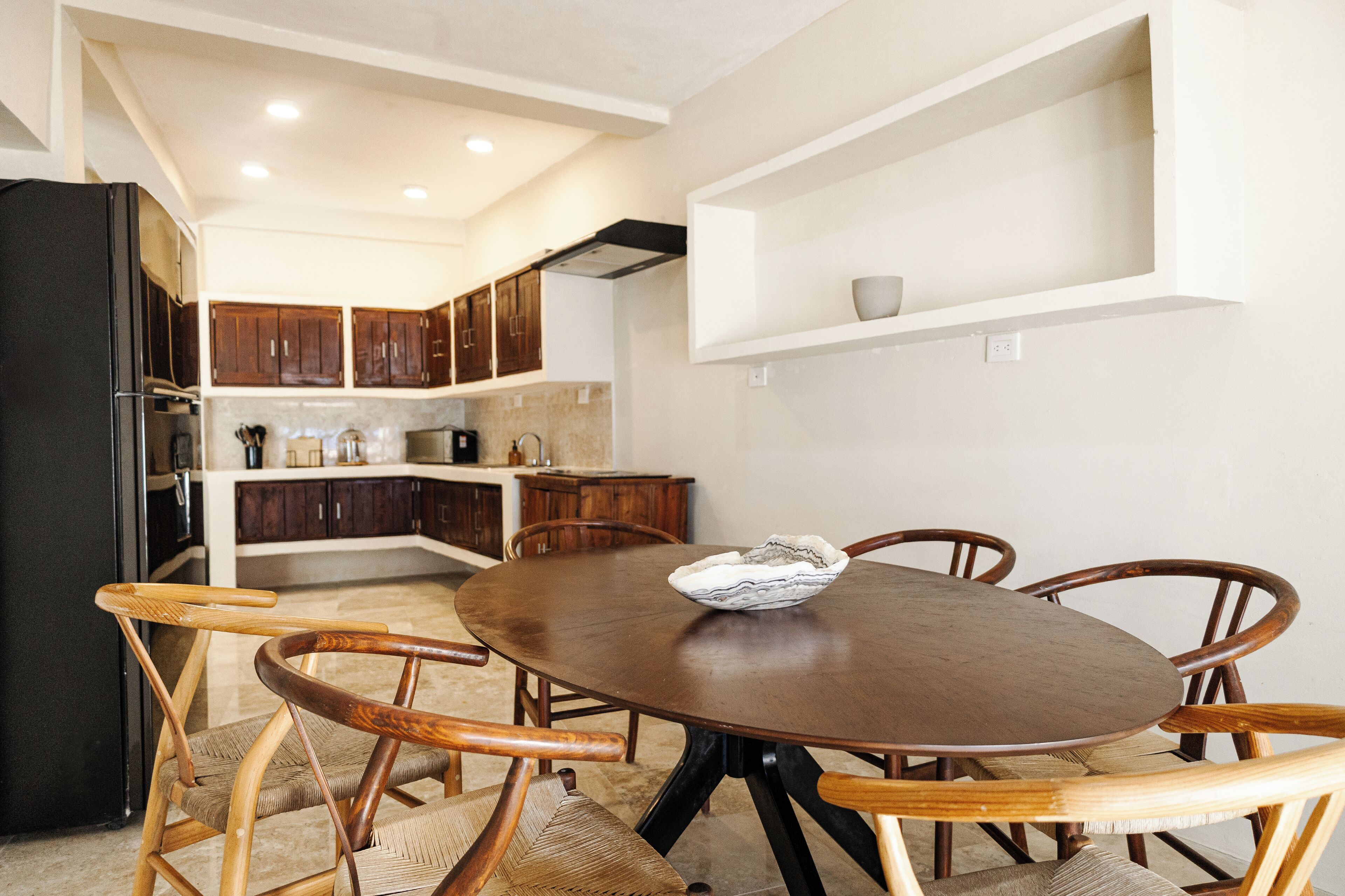 A dining area with a round wooden table and six chairs, a kitchen in the background, and a white shelf above the table.