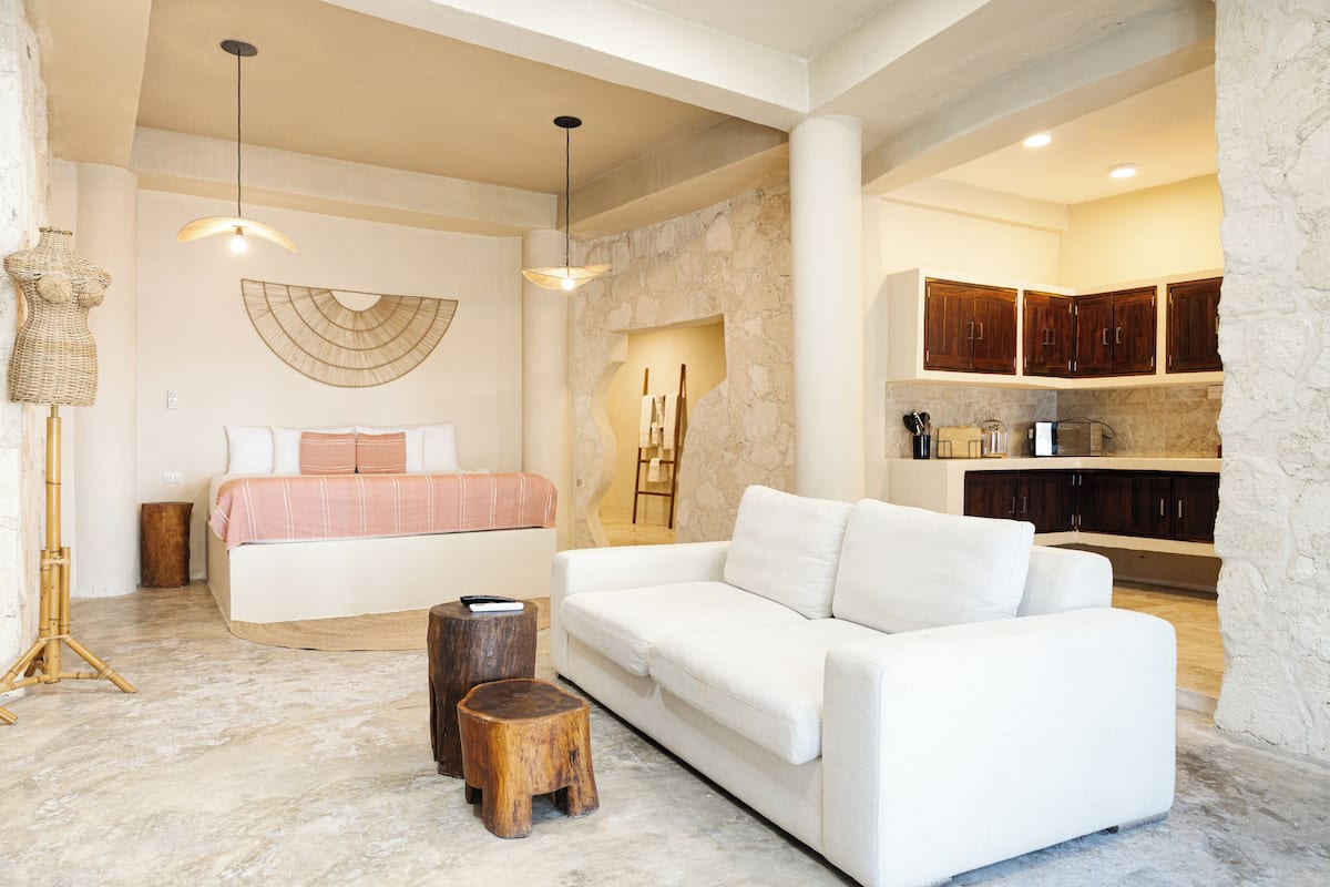 Un salón amplio con un sofá blanco, una cama con una colcha a rayas rosa y blanca, un taburete de madera y una obra de arte grande colgada en la pared.
