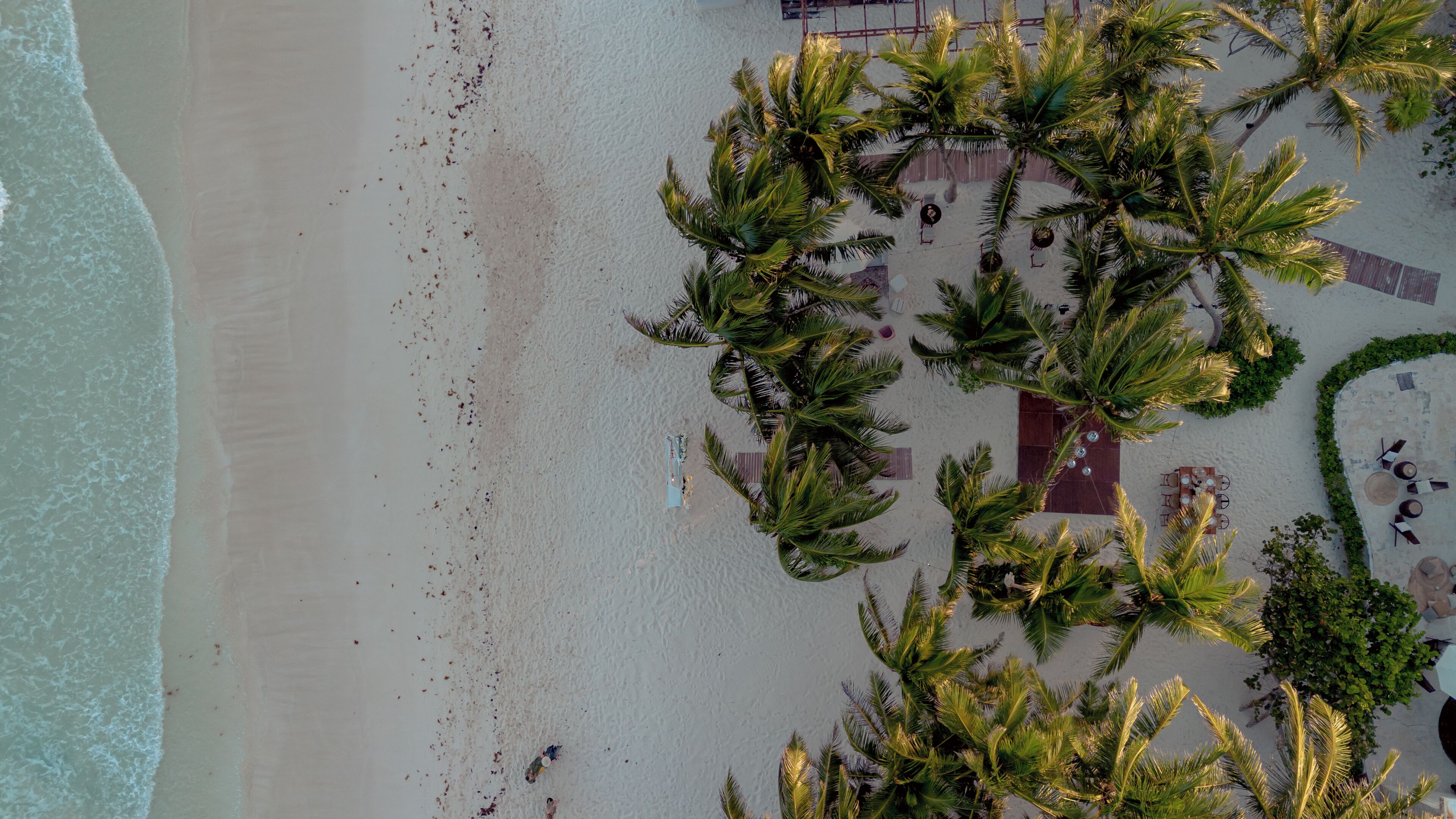 Luchtbeeld van een strand met palmbomen, enkele mensen en een klein gebouw met terras.