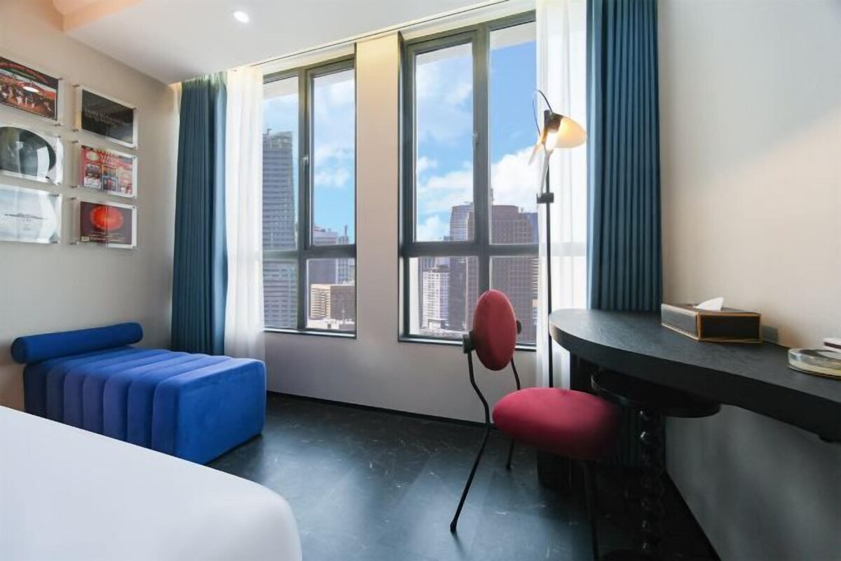 A modern hotel room with a large window, a blue sofa, a red chair, and a desk with a lamp.