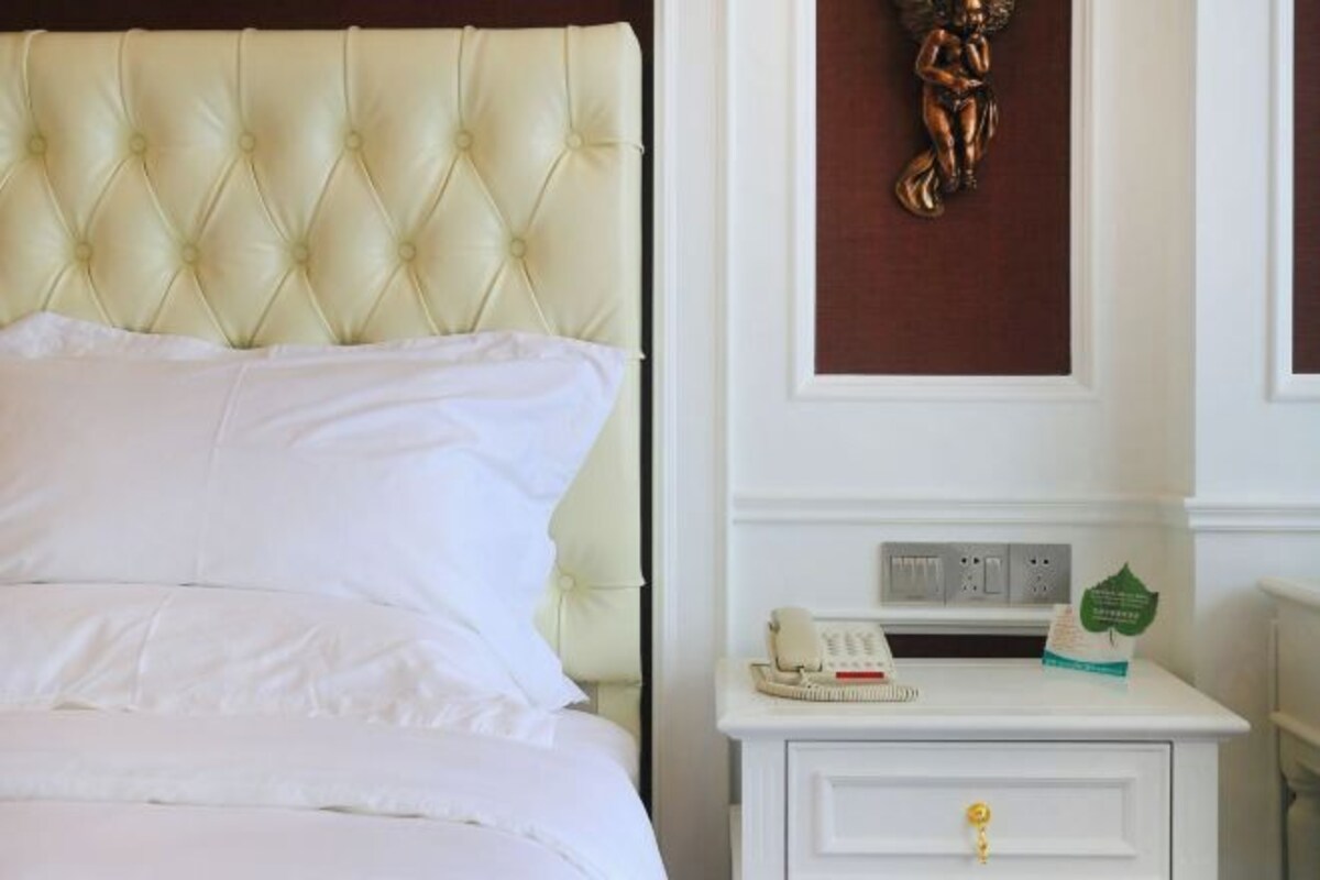 A neatly made bed with white linens and a tufted headboard, a white nightstand with a telephone and a green item on it, and a decorative angel on the wall.