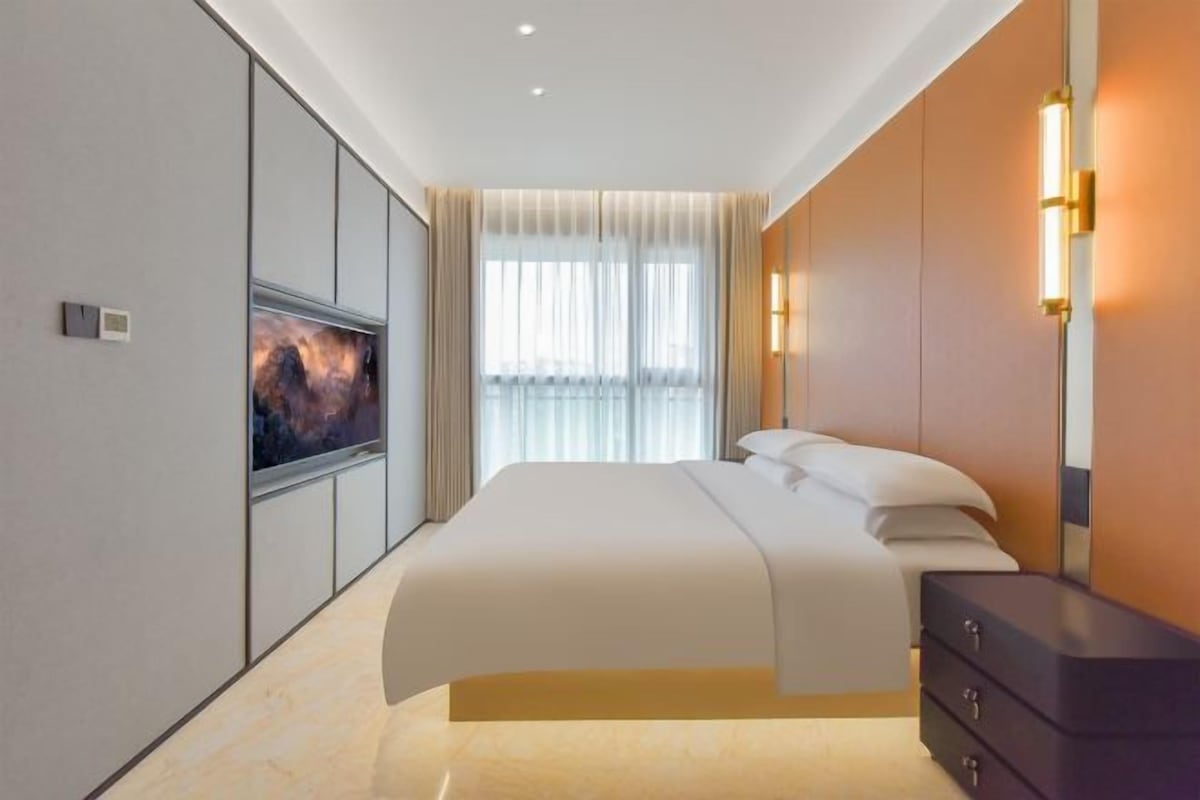 A modern hotel room with a large bed, a flat-screen TV, a dresser, and a large window with curtains.