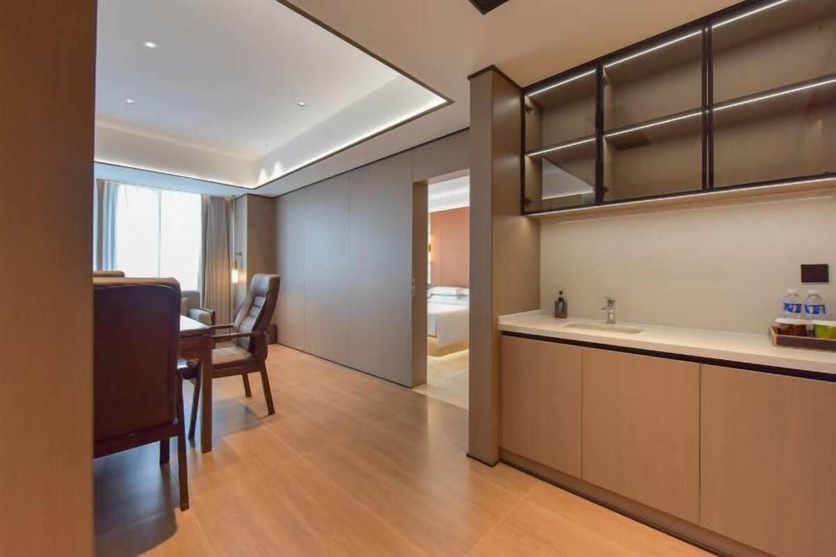 A modern hotel room with a dining area, a kitchen with a built-in shelf, and a large window providing natural light.