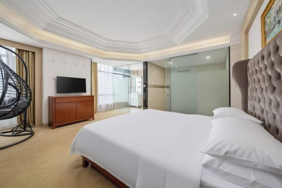 Quarto de hotel com cama, televisão, armário de madeira, divisória de vidro e cabeceira estofada.