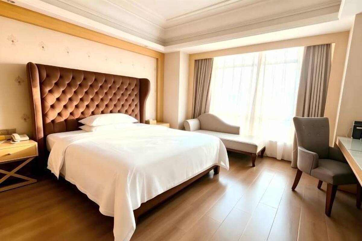 Quarto de hotel com uma cama grande, uma escrivaninha, uma cadeira e uma janela com cortinas.