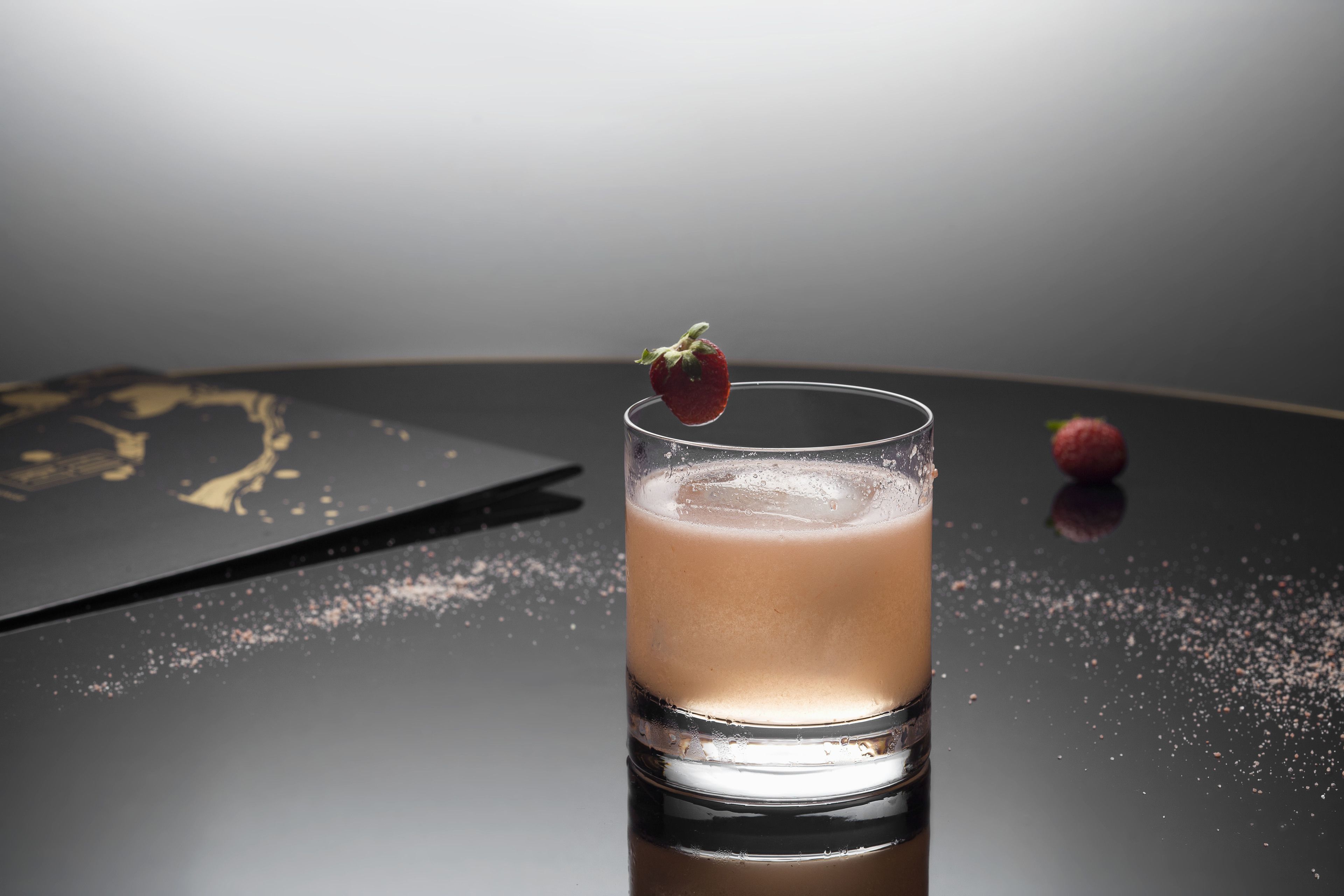 A glass of a light-colored beverage with a strawberry garnish on a reflective surface.