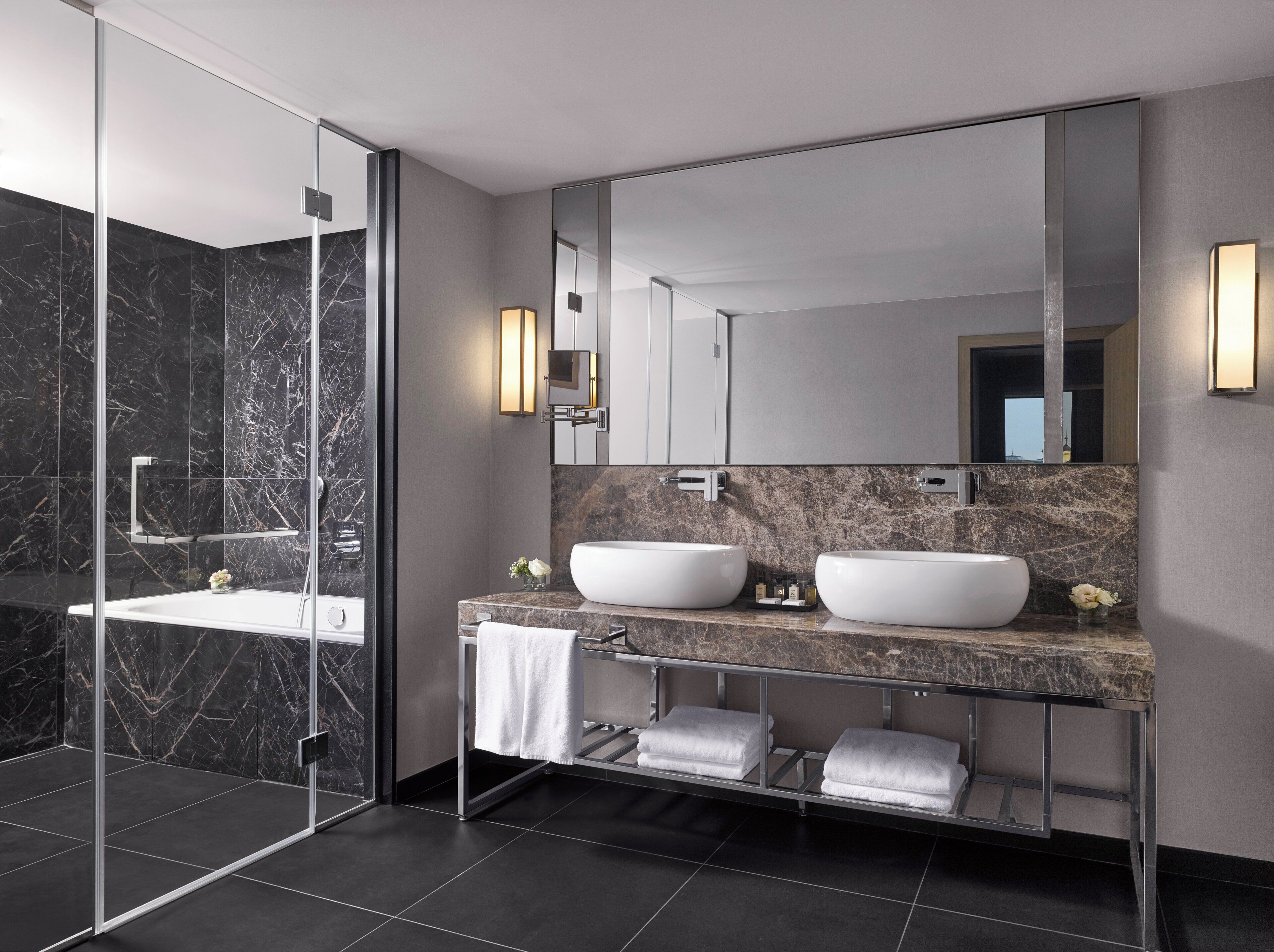 Un baño moderno con dos lavamanos, cabina de ducha con mampara de vidrio y zona de ducha con azulejos de mármol.