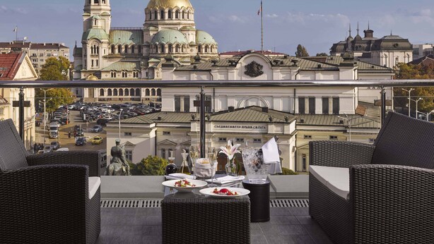 A rooftop terrace with wicker furniture, a dining table set with plates and glasses, and a view of a historic building with domes and a clock tower. - InterContinental Sofia by IHG (Sofia)