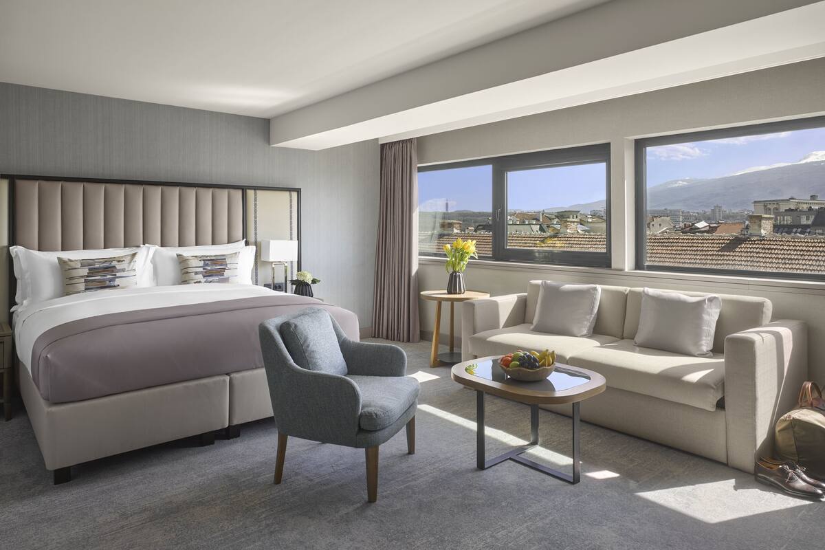 A modern hotel room with a large bed, a sofa, a chair, and a small table with a fruit bowl.