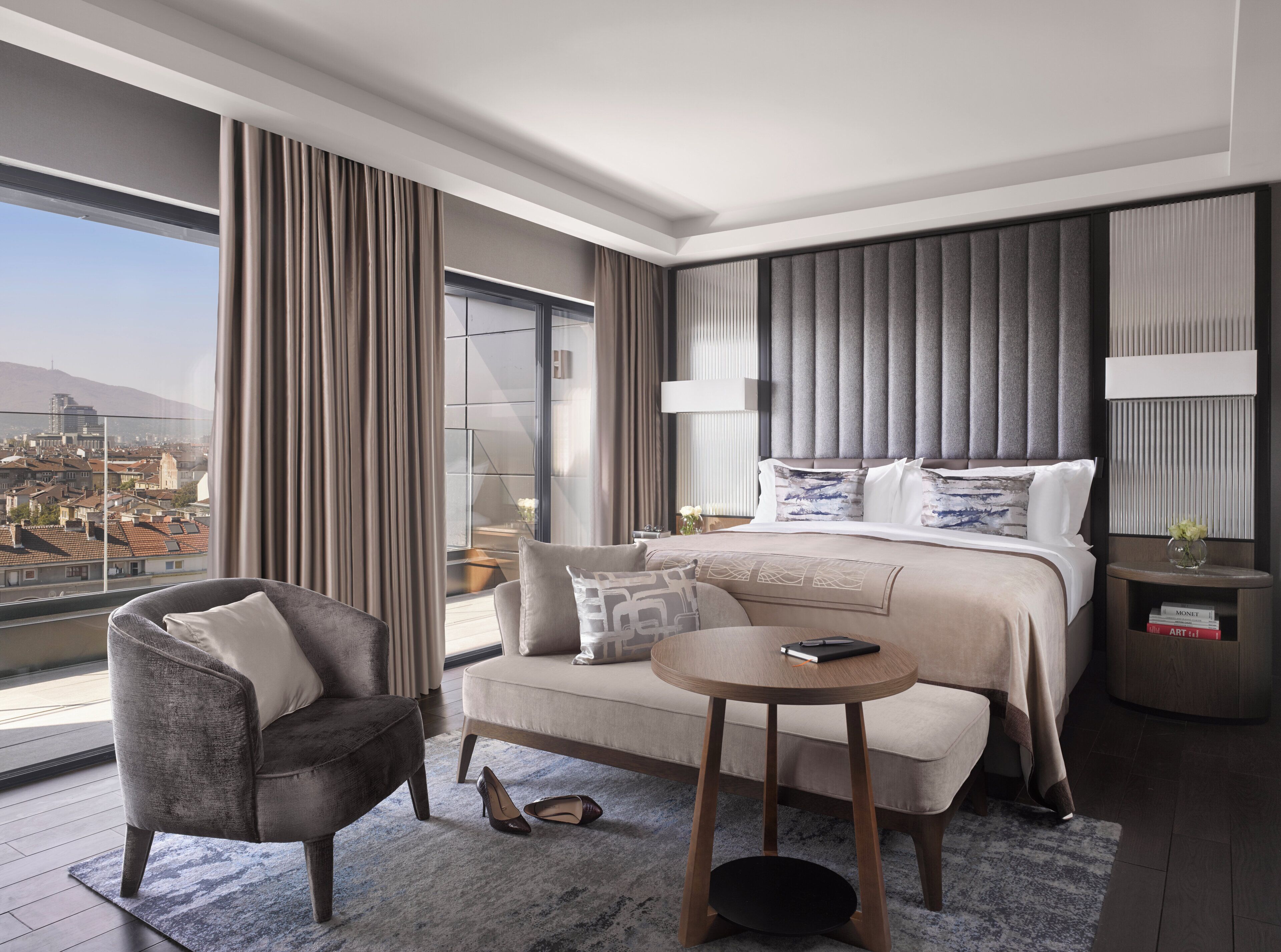 A modern hotel room with a large bed, a seating area with a chair and a small table, and a view of a cityscape through large windows.