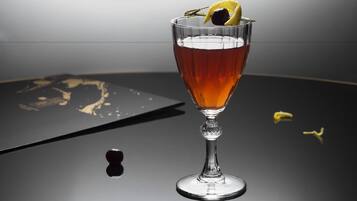 A glass of amber-colored beverage with a lemon slice and a cherry on a reflective surface.