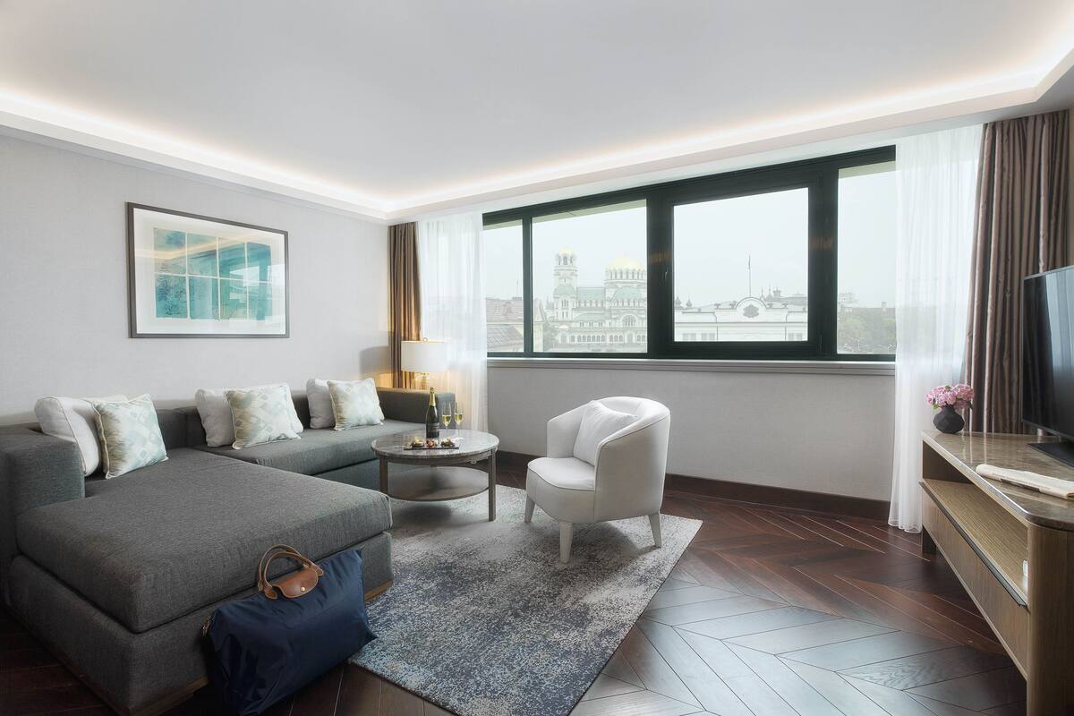 A modern hotel room with a grey sofa, a white armchair, a small round table with a lamp, and a TV on a wooden console.