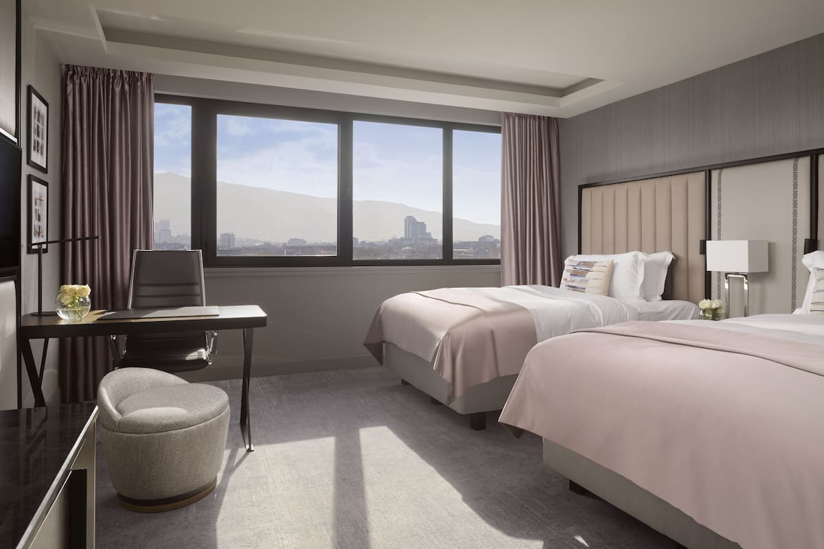 A hotel room with two beds, a desk with a chair, a small ottoman, and a large window offering a city view.