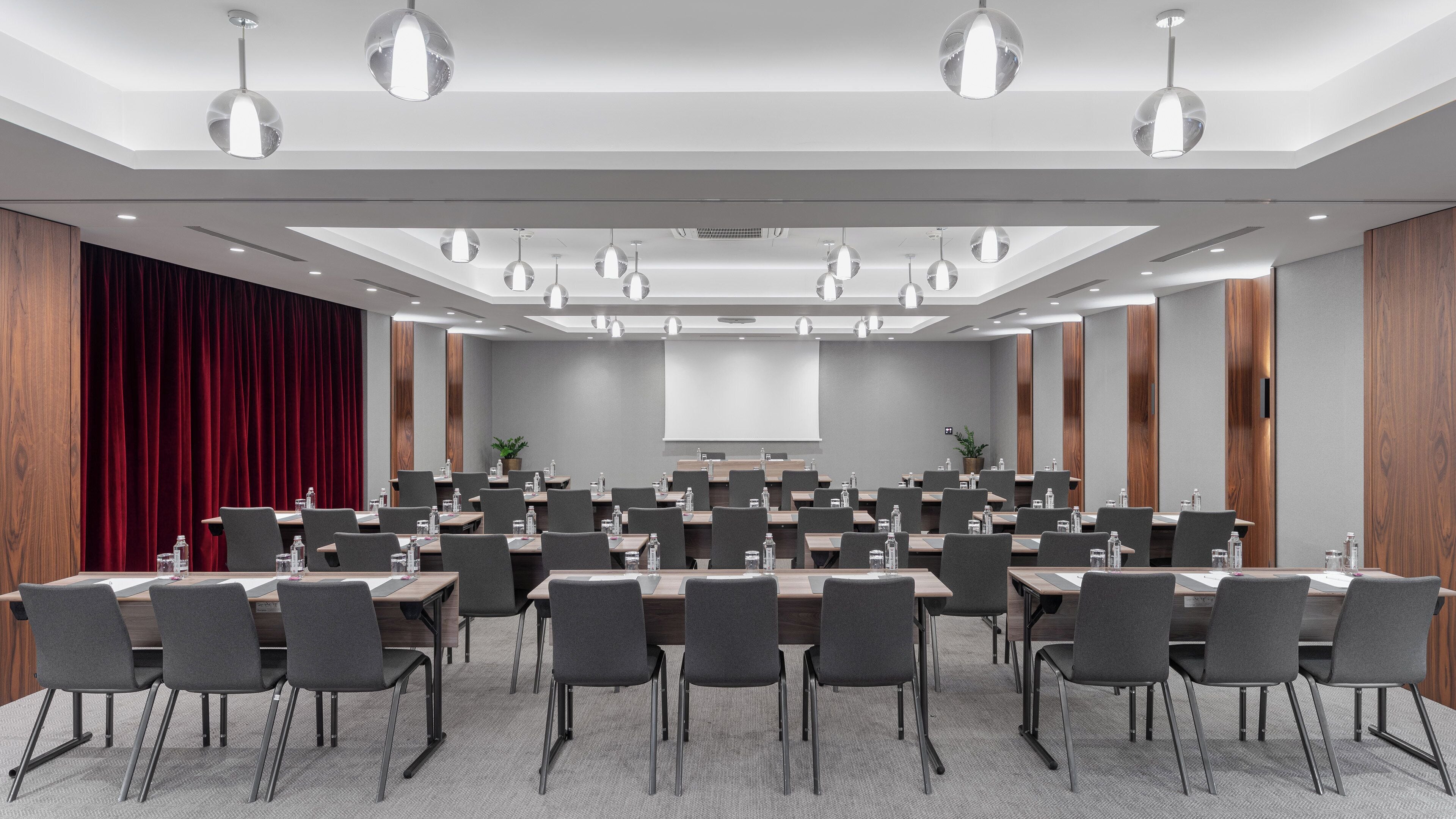 A conference room with multiple tables and chairs, a large screen, and overhead lighting.