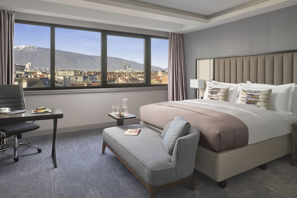 A hotel room with a large bed, a desk with a chair, a small table with a lamp, and a view of a cityscape with mountains.
