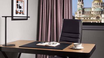 Uma cadeira de escritório moderna, uma mesa com um laptop, um par de óculos e uma chávena de café sobre uma mesa, com vista para um edifício com cúpulas através de uma janela.