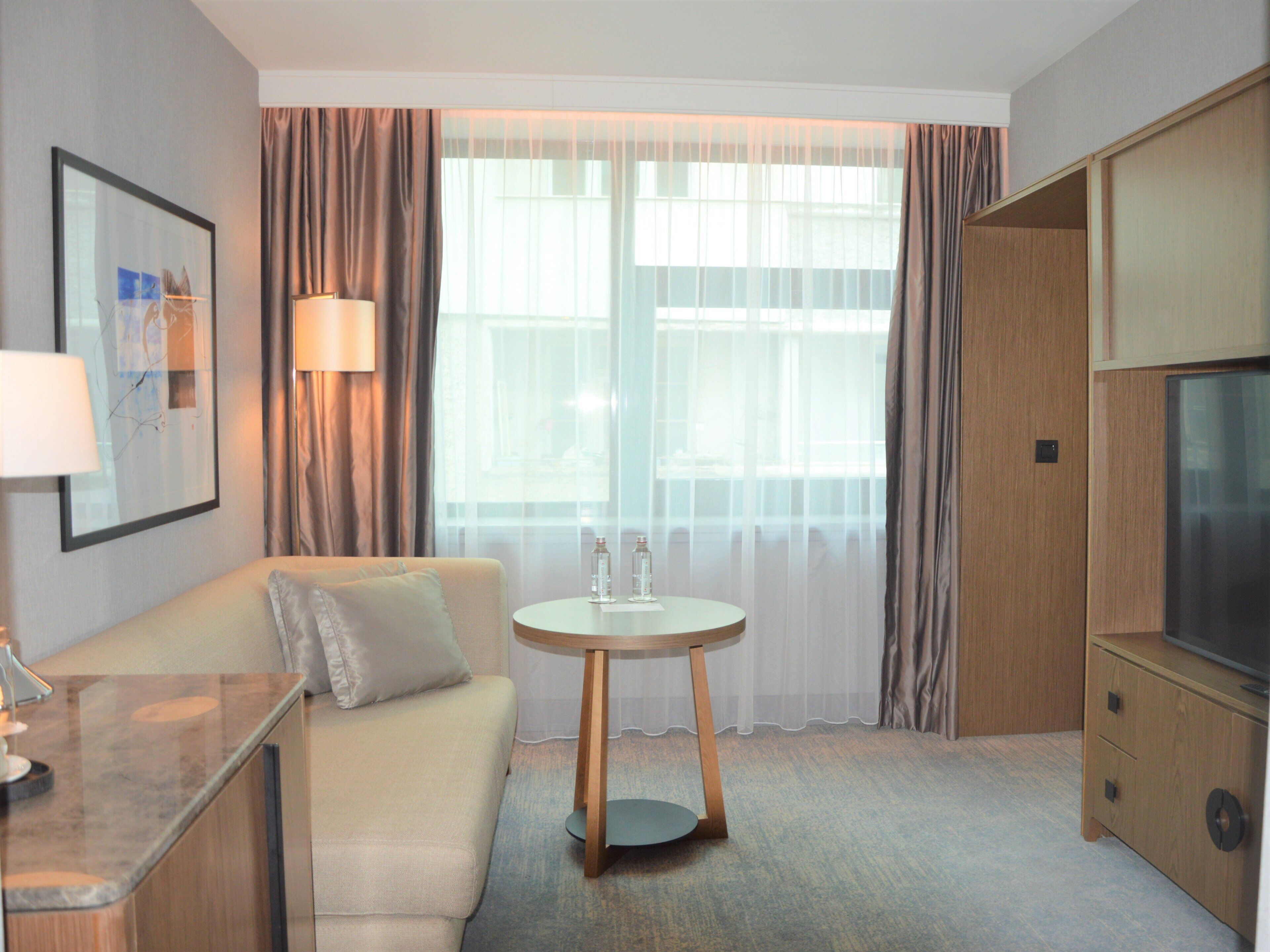A hotel room with a sofa, a small round table, a lamp, a TV, and a large window with curtains.