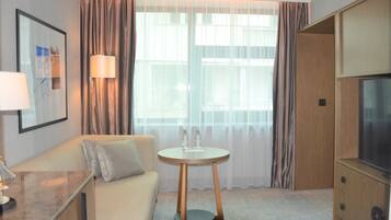 A hotel room with a sofa, a small round table, a lamp, a TV, and a large window with curtains.