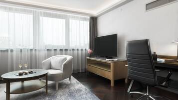 A modern hotel room with a large window, a flat-screen TV, a comfortable sofa, a desk with a chair, and a small round table with a tray of snacks.