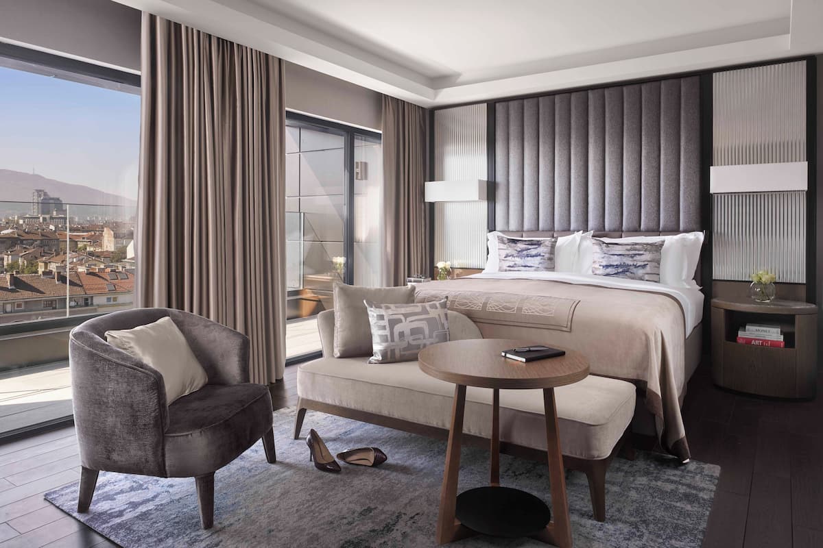 A modern hotel room with a large bed, a sofa, a small round table, and a view of a cityscape.