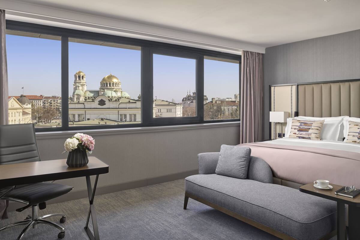 A hotel room with a large window, a bed with a headboard, a desk with a chair, and a small table with a cup and glasses.