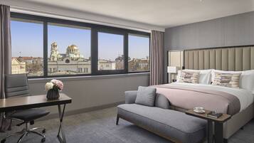 A hotel room with a large window, a bed with a headboard, a desk with a chair, and a small table with a cup and glasses.