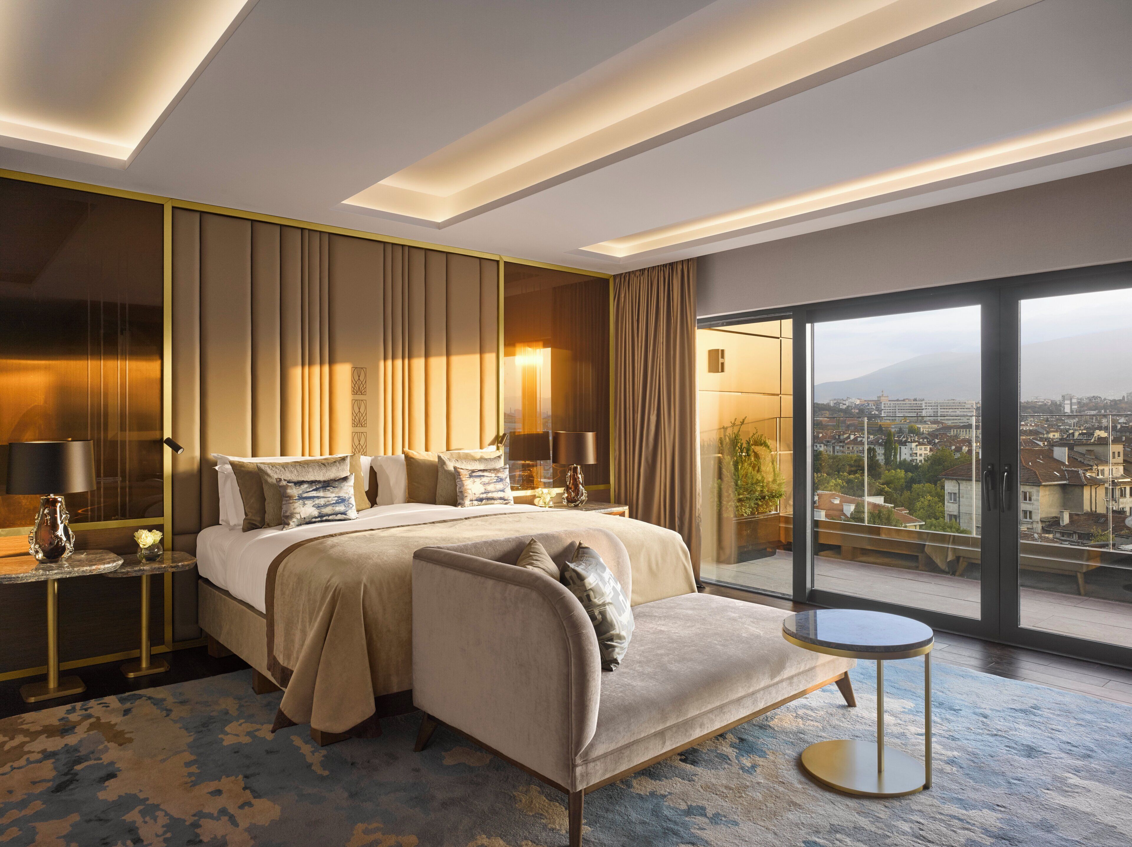 A modern hotel room with a large bed, a sofa, a small table, and a view of the city through large windows.