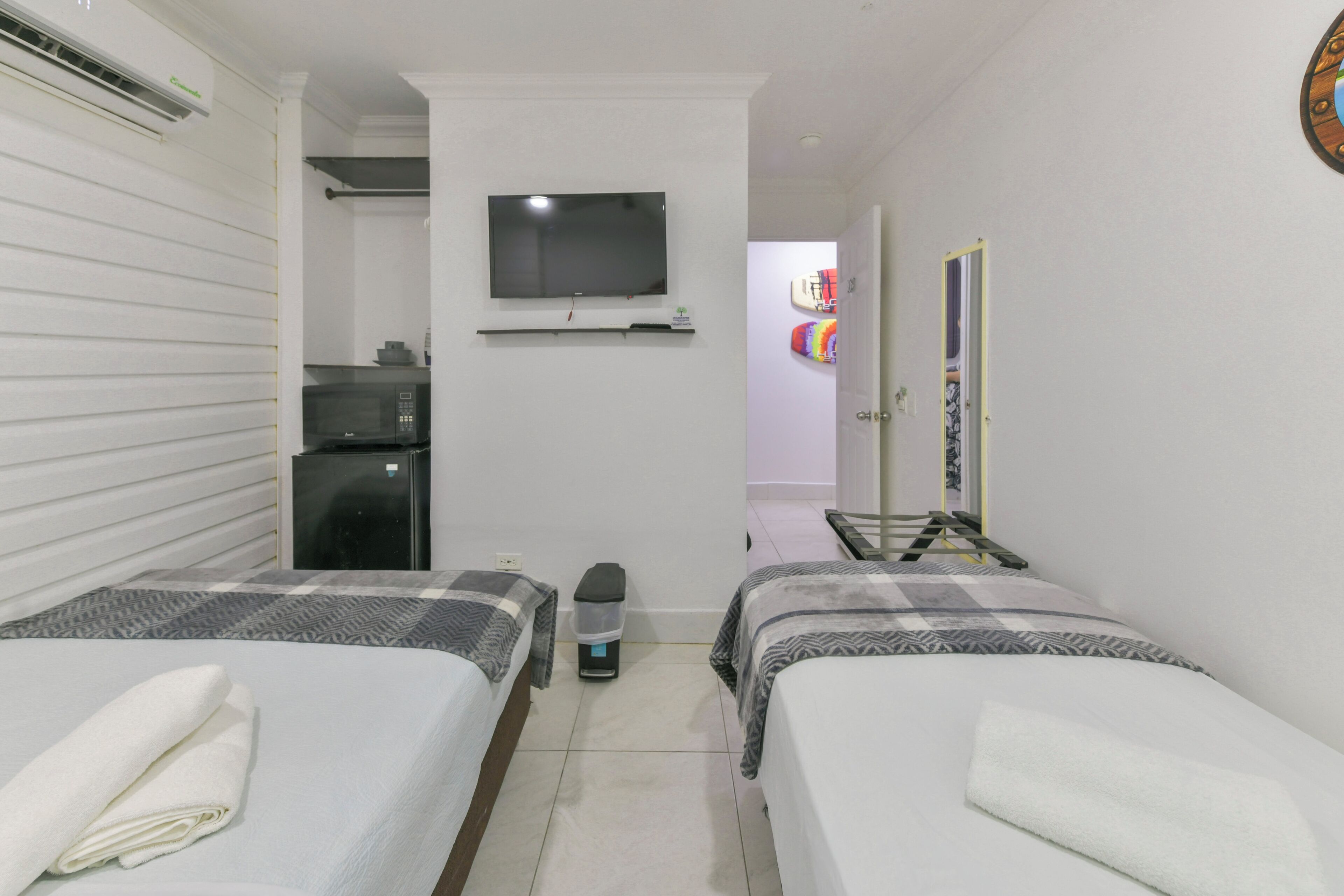 A hotel room with two beds, a TV mounted on the wall, a small kitchenette, and a door leading to another room.