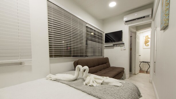 Un dormitorio con cama, un sofá marrón, un televisor en la pared y una ventana con persianas.