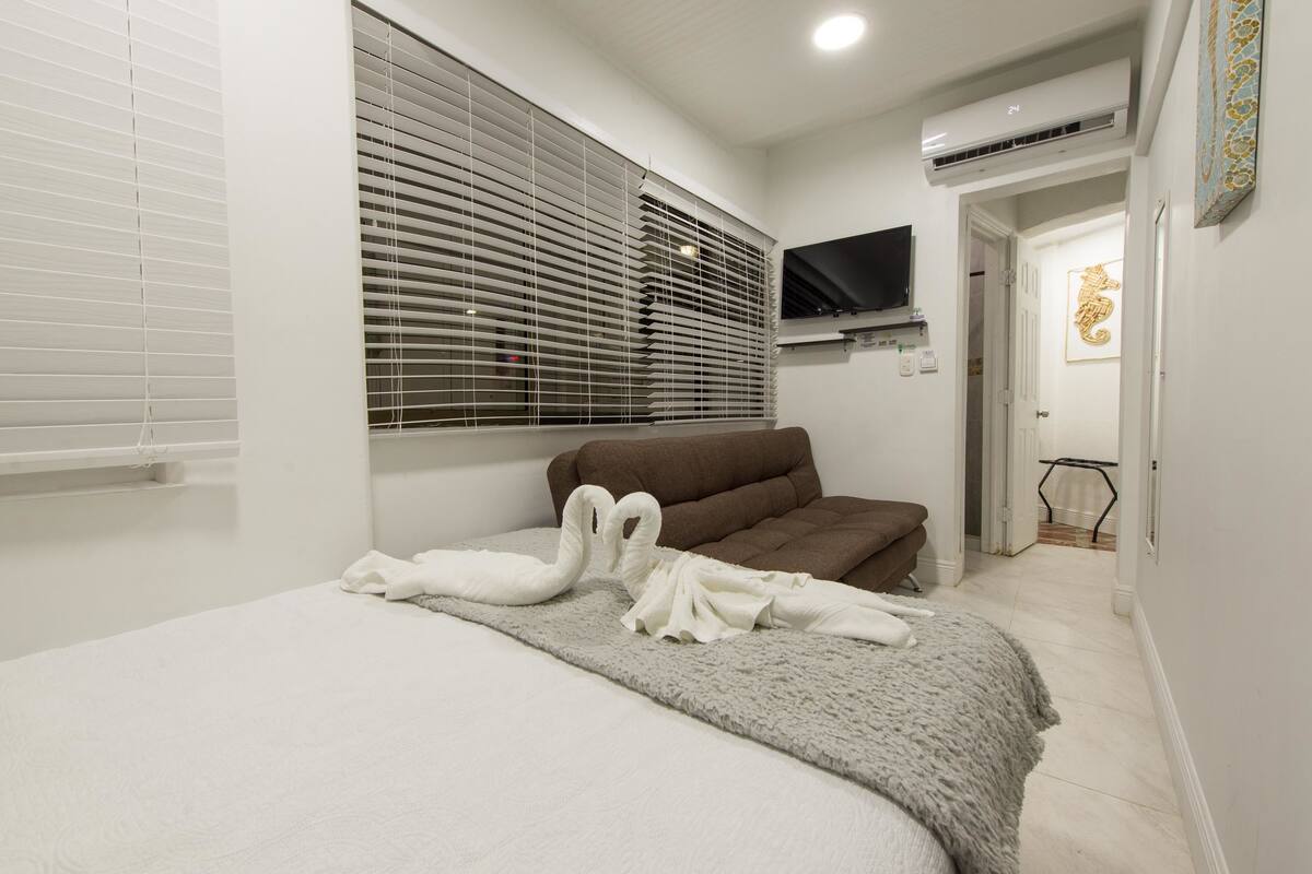 A bedroom with a bed, a brown sofa, a TV mounted on the wall, and a window with blinds.