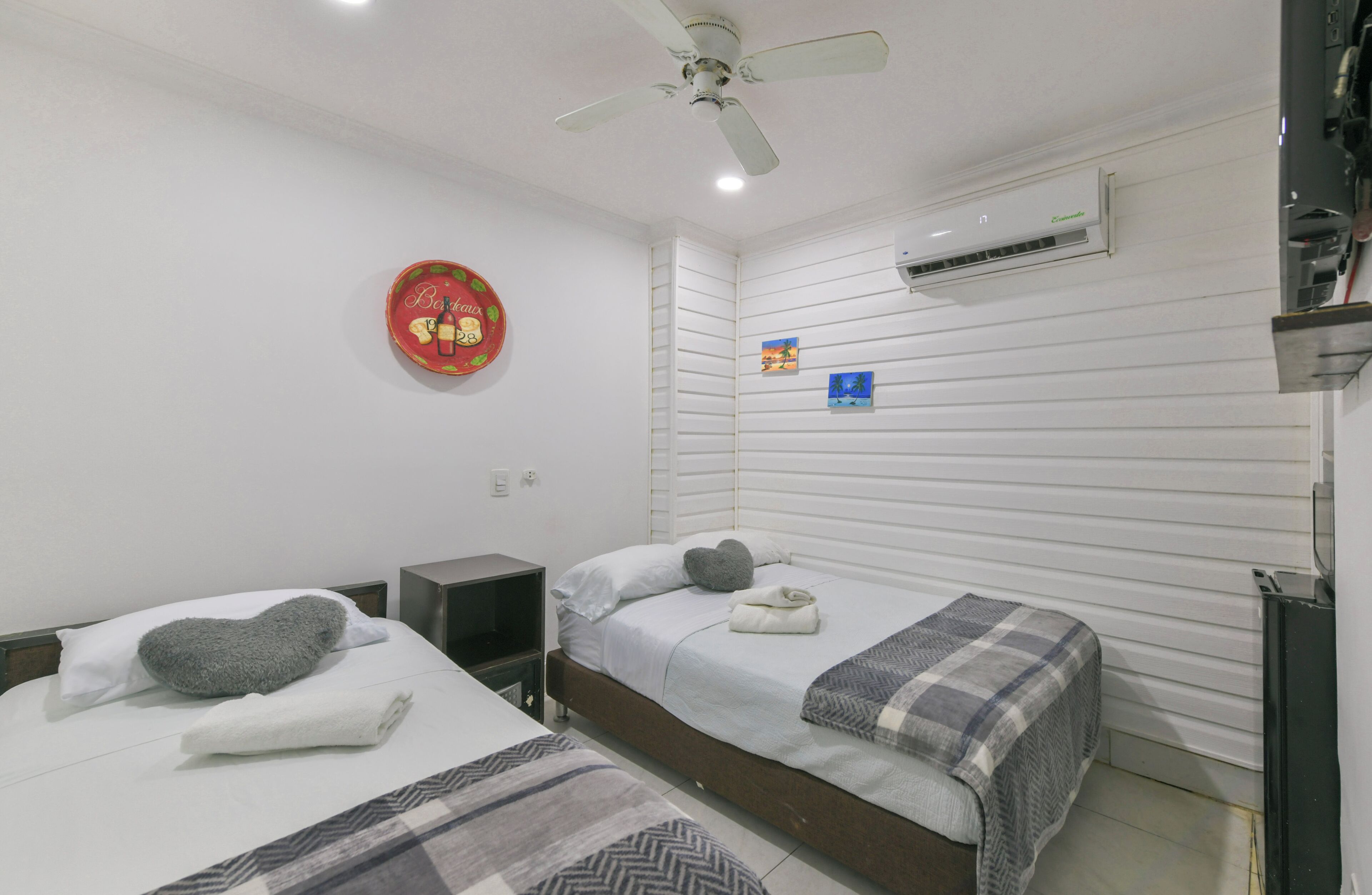 A hotel room with two beds, a ceiling fan, an air conditioner, a small table, and a wall decoration.