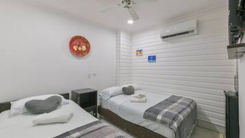 Quarto de hotel com duas camas, um ventilador de teto, ar condicionado, uma pequena mesa e uma decoração na parede.