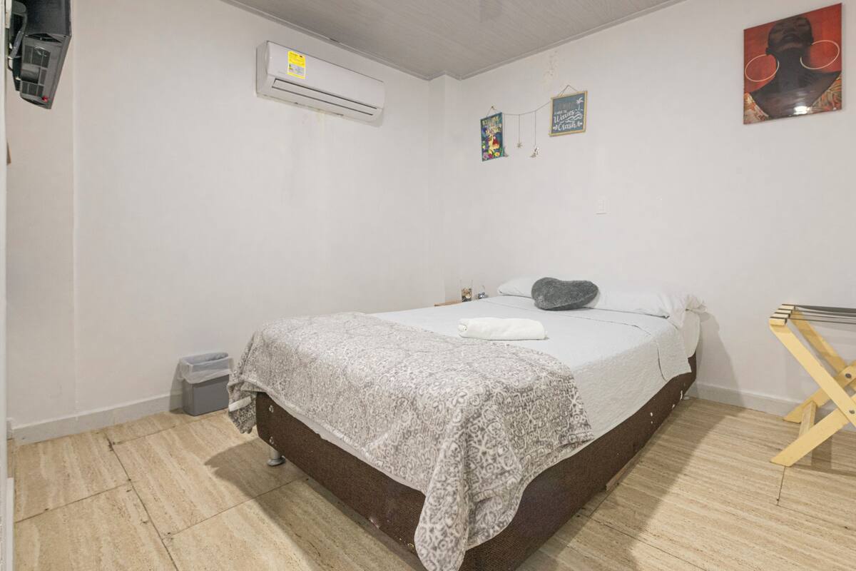A bedroom with a bed, a small bedside table, a wall-mounted air conditioner, a wall-mounted drying rack, and a small trash bin.
