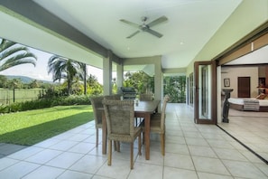 A spacious patio with a dining table and chairs, a ceiling fan, and a view of a lush garden and mountains.