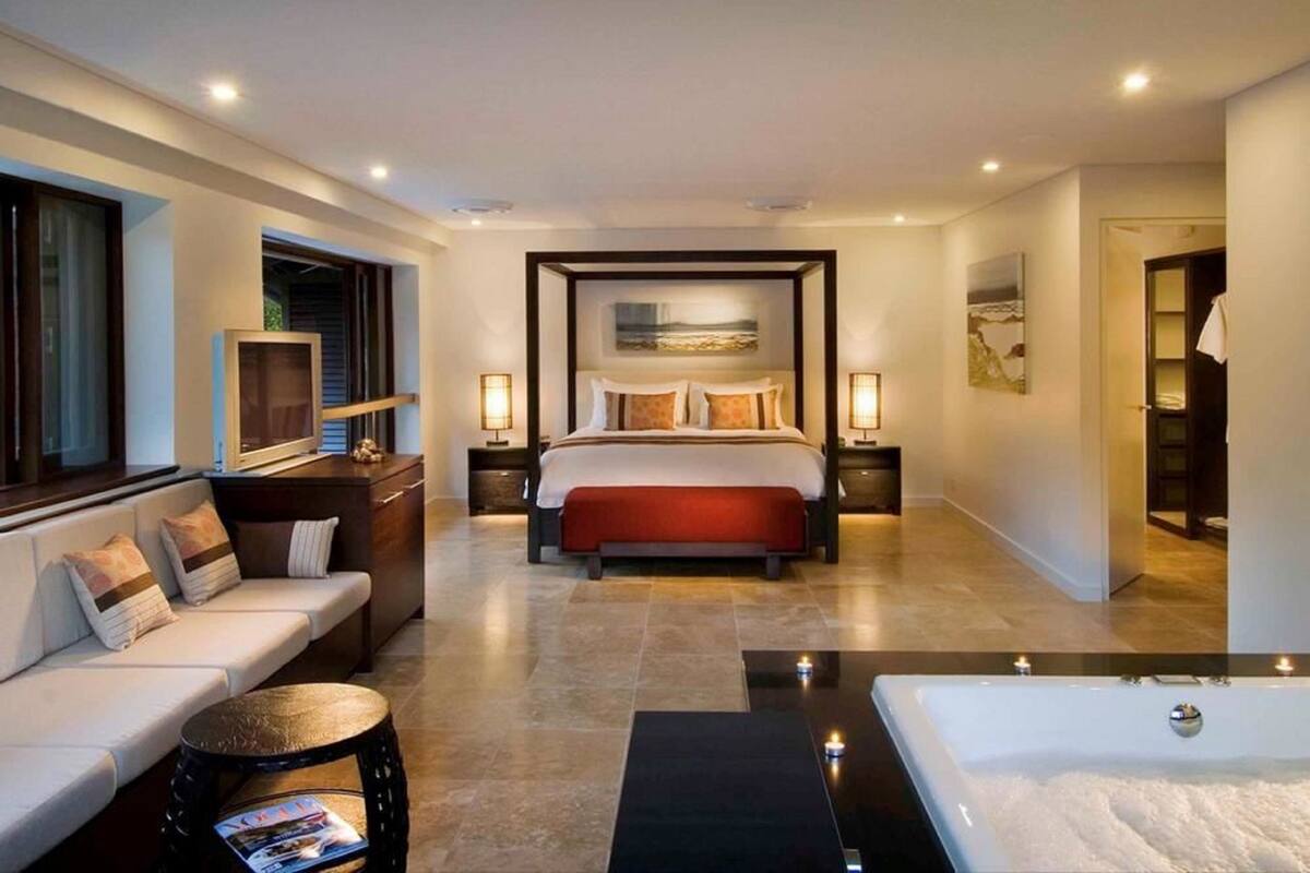 A modern hotel room with a large bed, a sitting area with a sofa and armchair, a small round table with a magazine, a bathtub, and a hallway leading to another room.