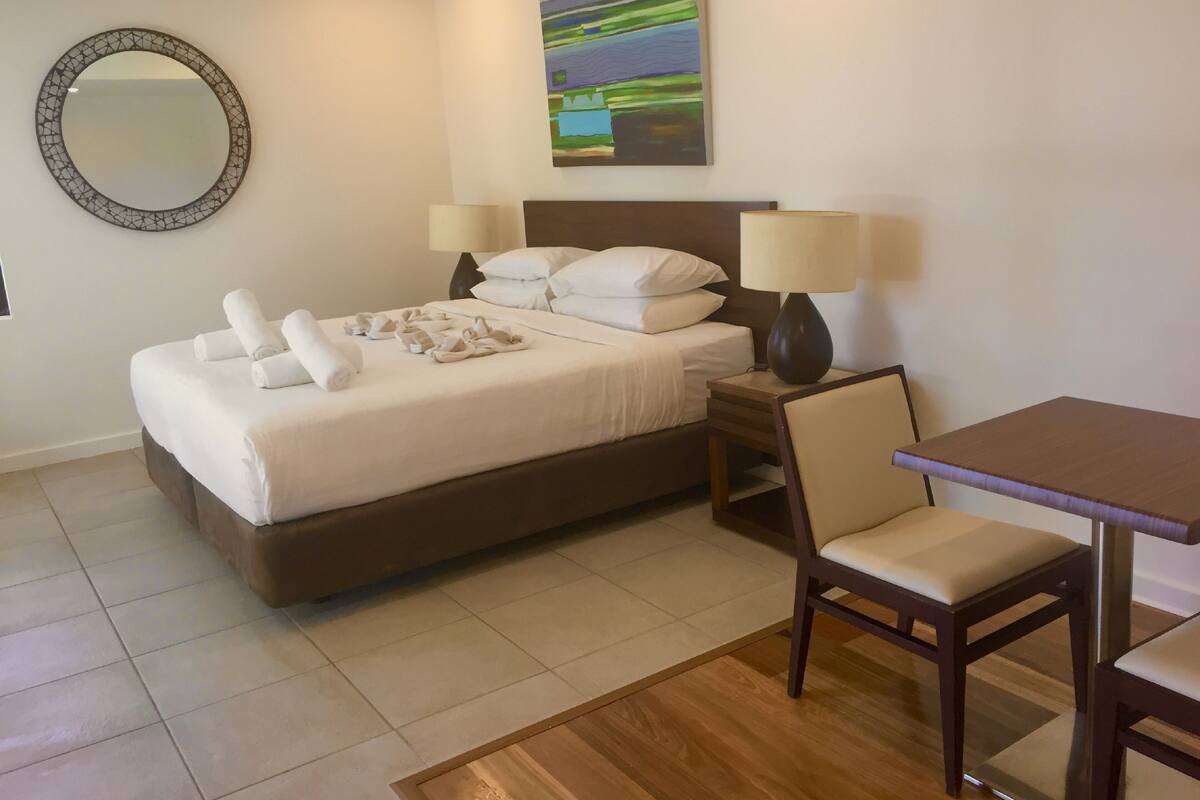 A neatly made bed with towels, a bedside table with a lamp, a chair, and a painting on the wall.
