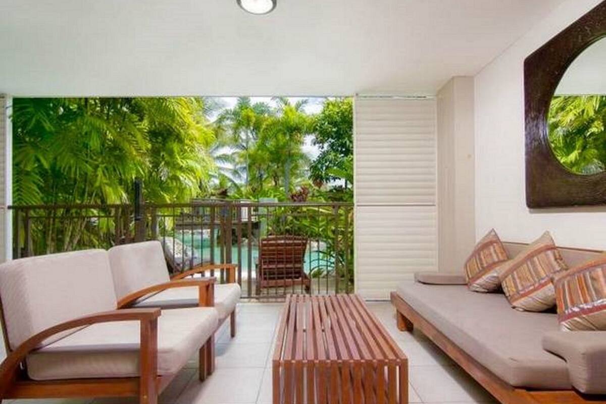 A balcony with a view of lush greenery and a pool. There is a sofa and a wooden coffee table on the balcony.