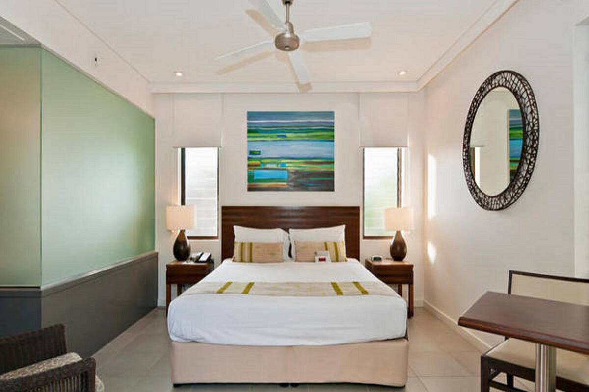 A hotel room with a large bed, two bedside tables, a ceiling fan, a painting, a mirror, and a desk.
