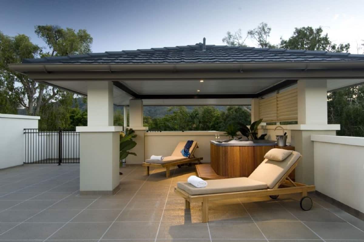 A covered outdoor area with two lounge chairs, a small wooden table, and a hot tub.