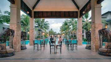 A covered outdoor seating area with a pool, stone pillars, and palm trees.