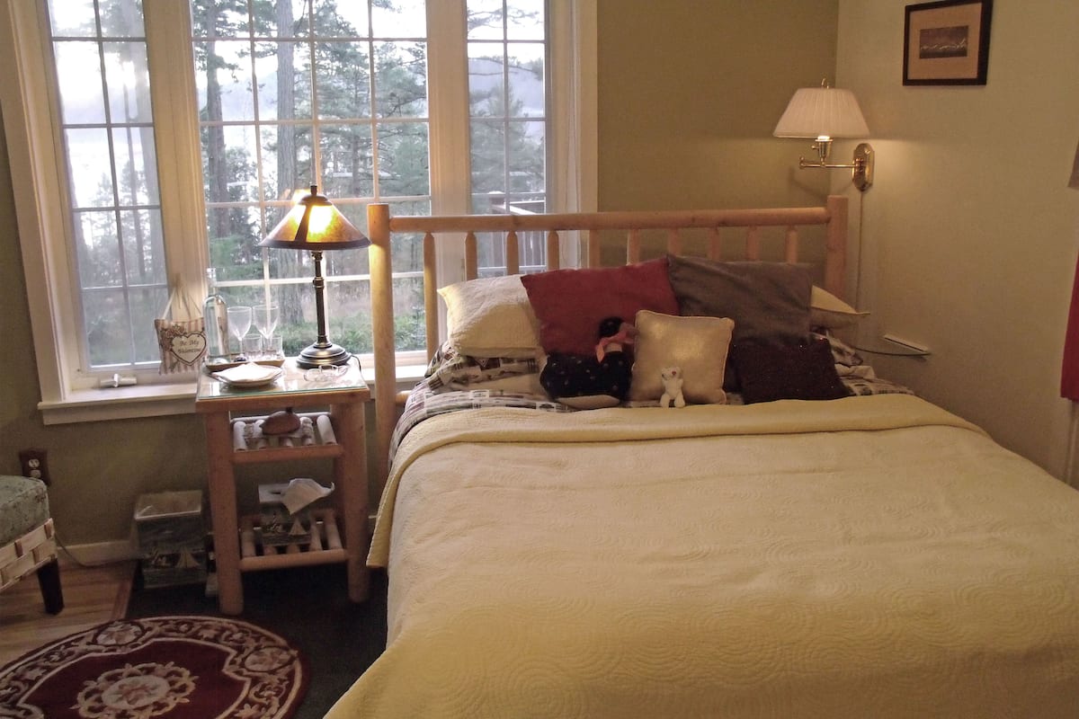 A cozy bedroom with a wooden bed, a bedside table with a lamp, a window with a view of trees, and a small rug on the floor.