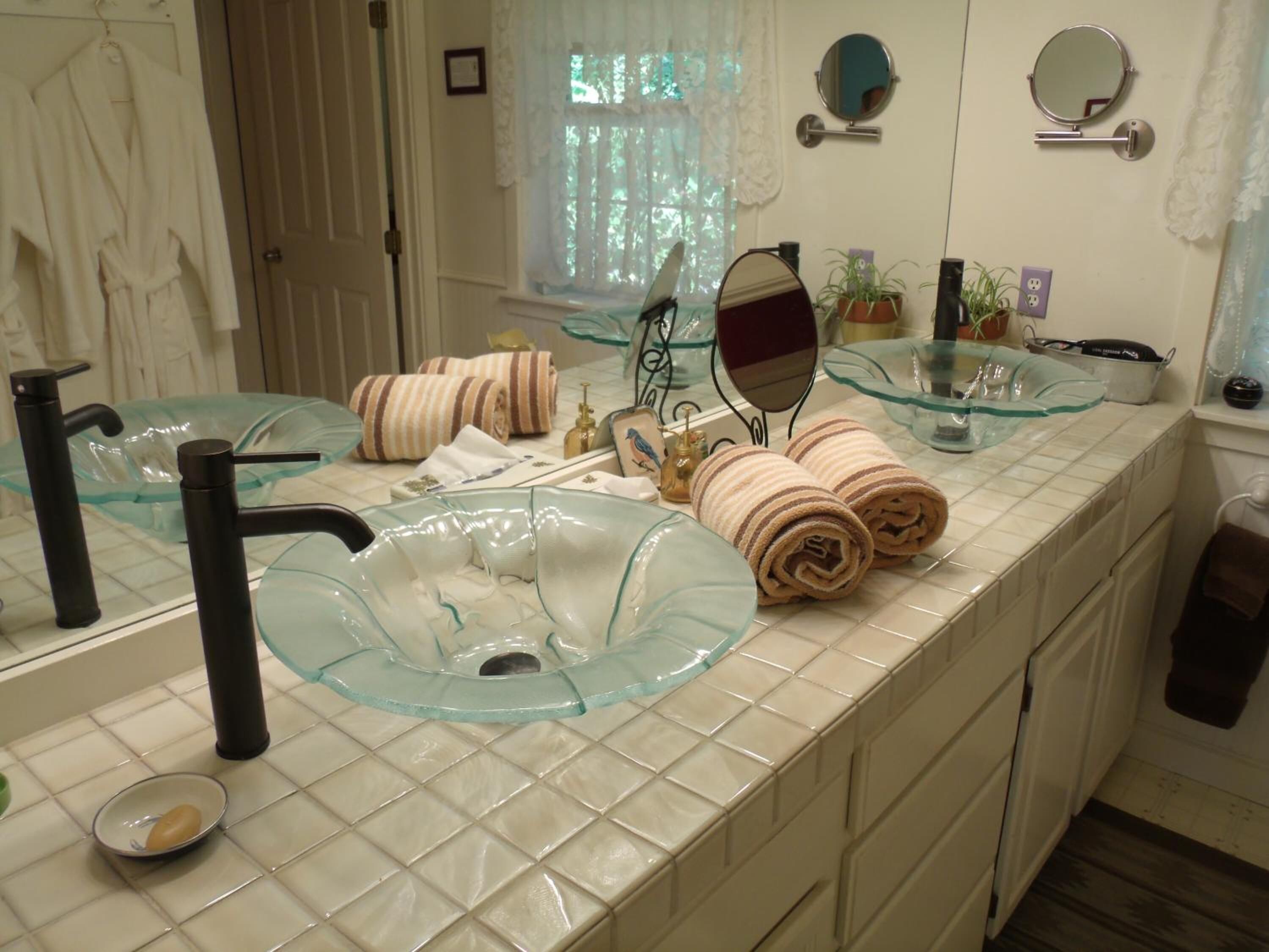 A bathroom with a glass sink, two mirrors, a towel rack with towels, a potted plant, and a window with curtains.