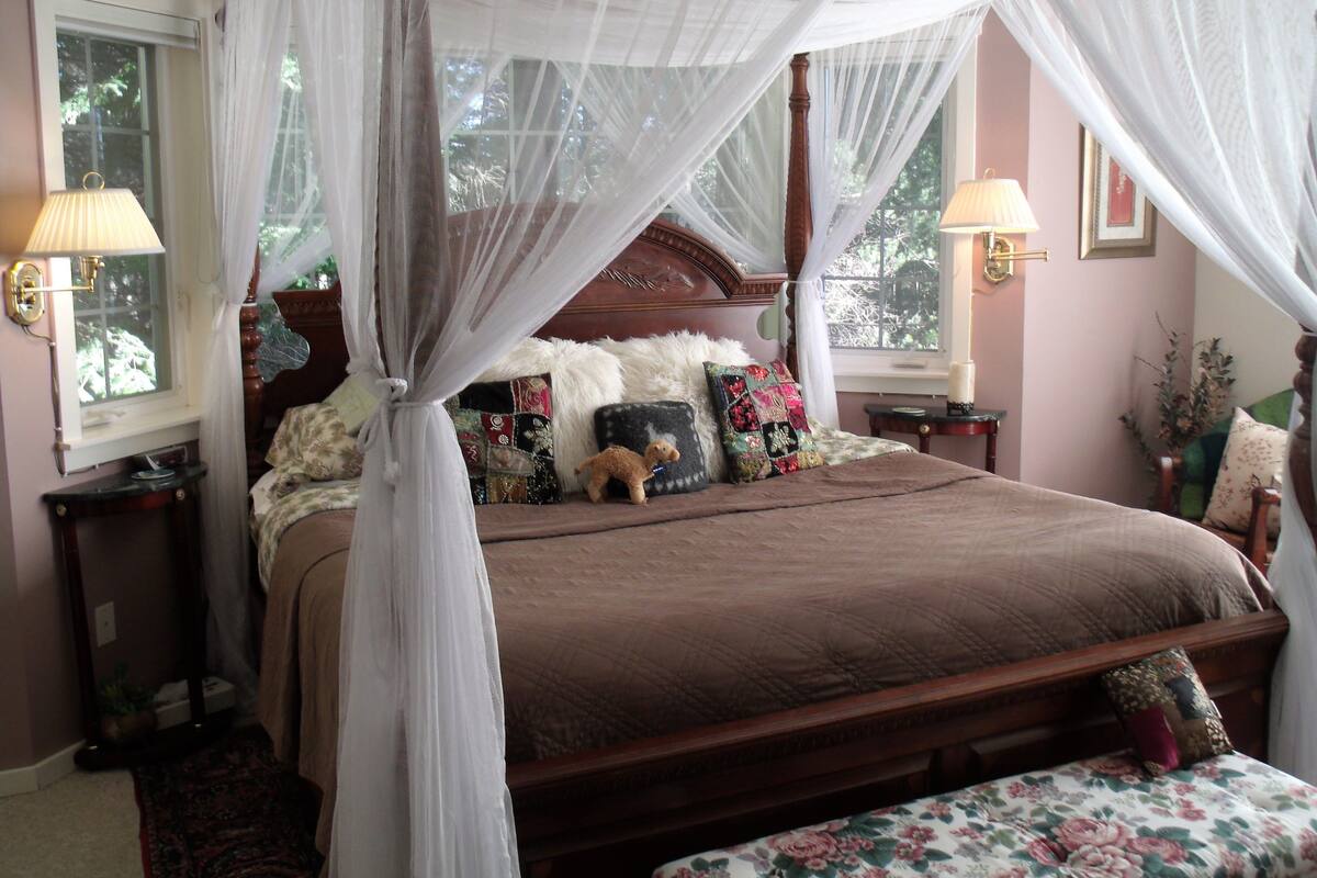 A four-poster canopy bed with a brown bedspread, multiple decorative pillows, and a floral-patterned bench at the foot of the bed.