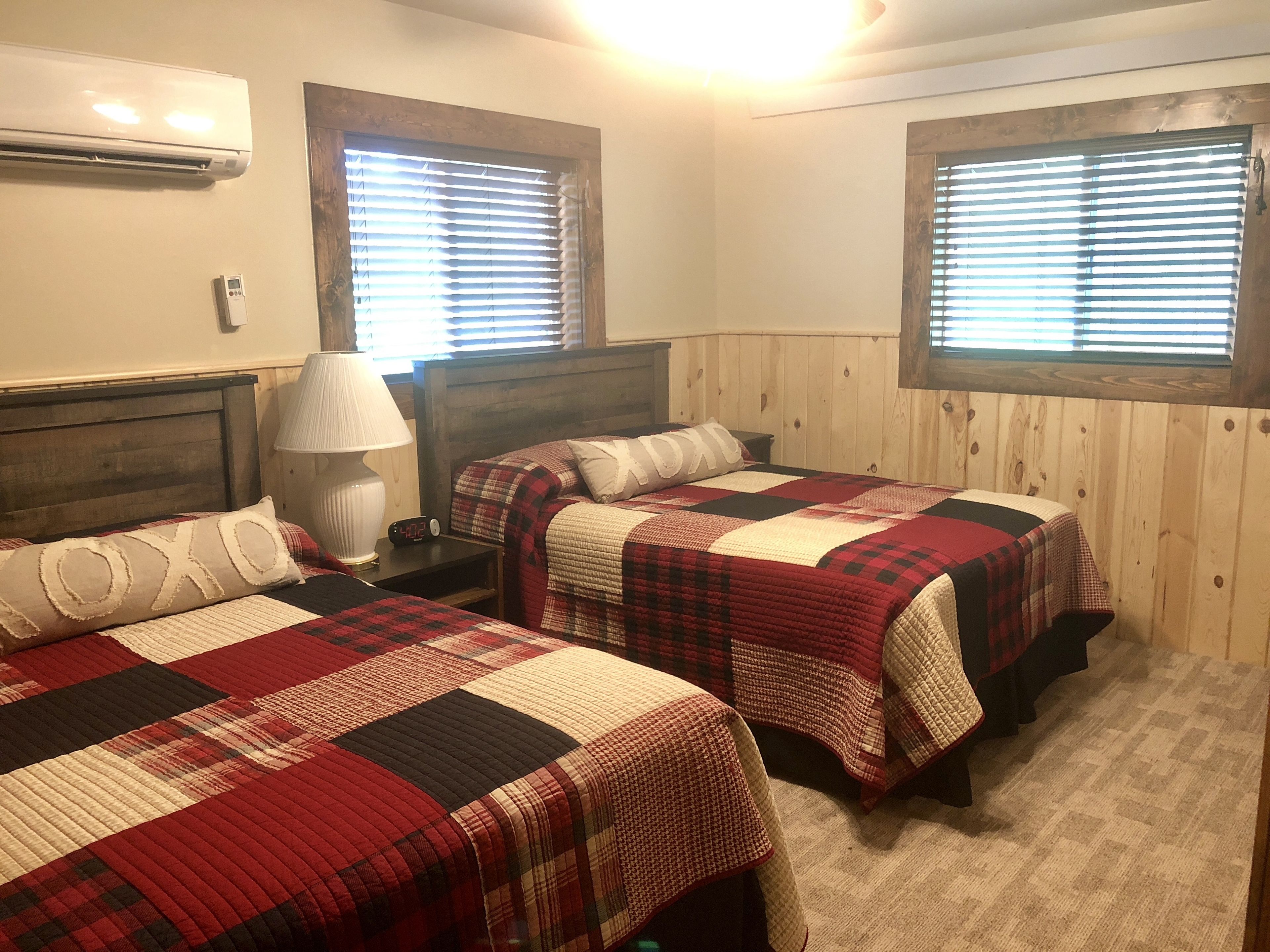 Une chambre avec deux lits, chacun recouvert d’une courtepointe à carreaux rouges et noirs, une table de chevet avec une lampe et un climatiseur fixé au mur.